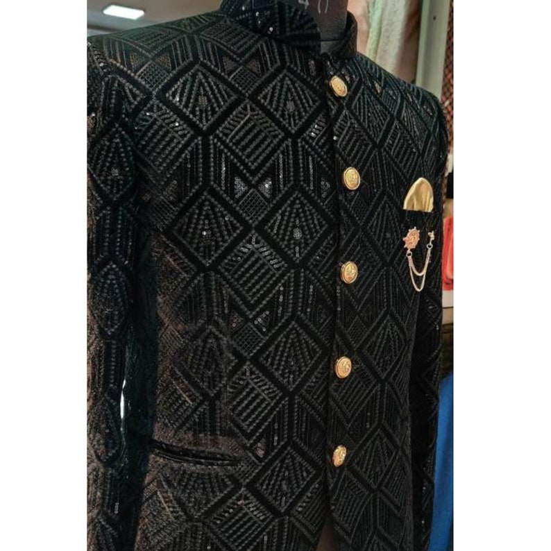 May include: A black velvet blazer with a geometric pattern and gold buttons. The blazer has a mandarin collar and a pocket on the left side. A gold chain with a decorative pin is attached to the pocket.