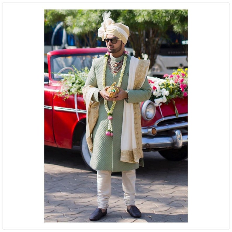 May include: A man wearing a sage green sherwani with a white and gold embroidered shawl, a gold necklace, and a gold turban. He is holding a gold box and standing in front of a red car.