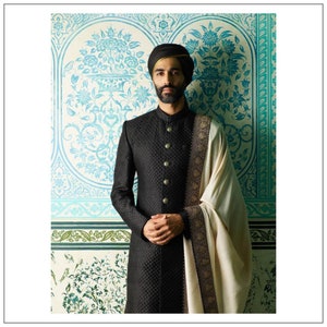 May include: A man wearing a black quilted long coat with gold buttons and a cream-colored shawl with a black and gold border. He is also wearing a turban.