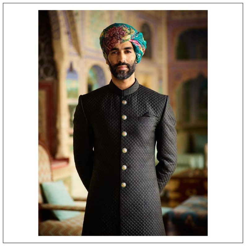 May include: A man wearing a black quilted jacket with gold buttons and a colorful turban. He is standing in a room with ornate walls and a couch in the background.