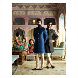 May include: Two men in traditional Indian clothing stand in a room with ornate walls and a patterned rug. The man on the left is wearing a blue long-sleeved jacket with gold buttons and white pants. The man on the right is wearing a black long-sleeved jacket with gold buttons and white pants. Both men are wearing turbans.
