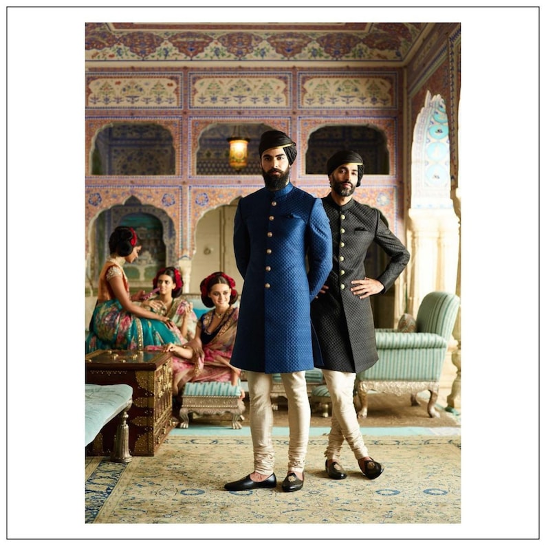 May include: Two men in traditional Indian clothing stand in a room with ornate walls and a patterned rug. The man on the left is wearing a blue long-sleeved jacket with gold buttons and white pants. The man on the right is wearing a black long-sleeved jacket with gold buttons and white pants. Both men are wearing turbans.