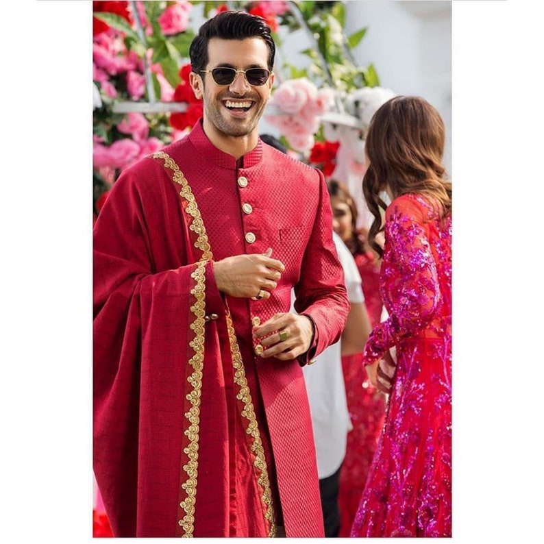 May include: A man wearing a red sherwani with gold embroidery and a matching red shawl. He is smiling and wearing sunglasses.