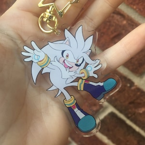 May include: A clear acrylic keychain featuring a cartoon depiction of Silver the Hedgehog from the Sonic the Hedgehog video game series. Silver is depicted with his signature white fur, red eyes, and blue and yellow bracelets. The keychain is attached to a gold star-shaped charm.