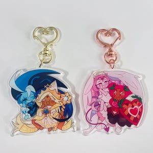 May include: Two acrylic keychains with cartoon character designs. One features characters in blue, gold, and white, and the other in pink, red, and white. Each keychain has a heart-shaped clasp.