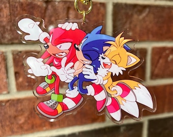 May include: A clear acrylic keychain featuring Sonic the Hedgehog, Tails, and Knuckles. The characters are drawn in a cartoon style and are shown hugging each other. Sonic is blue, Tails is yellow and orange, and Knuckles is red.