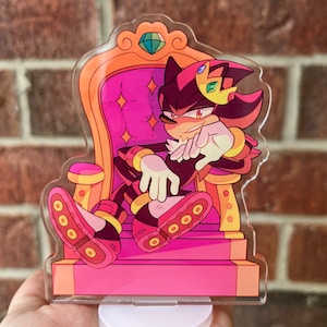 May include: A black and red cartoon character with a crown sits on a pink and gold throne. The character has a serious expression and is wearing a gold bracelet. The throne has a large, green gem on the top.