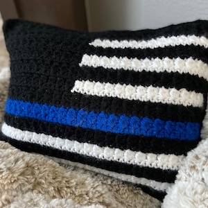 May include: A black and white crocheted pillow with a blue stripe across the middle. The pillow features a design resembling the American flag.