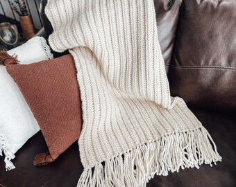 May include: A cream-coloured crocheted throw blanket with fringe on a brown leather sofa. The blanket is folded over a brown and white cushion.