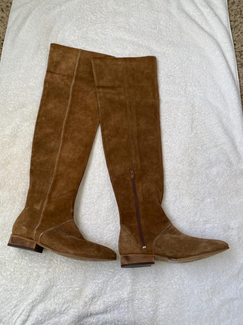 Pu&ograve; includere: Un paio di stivali al ginocchio in pelle scamosciata marrone con tacco piatto. Gli stivali hanno una cerniera laterale e una cucitura sul retro. Gli stivali sono su una superficie bianca.