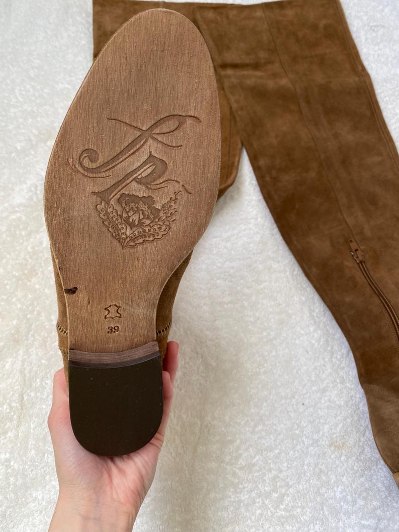 Pu&ograve; includere: Stivali al ginocchio in pelle scamosciata marrone. La suola presenta un logo decorativo e la taglia 39. Gli stivali hanno una cerniera laterale e un tacco largo. Gli stivali sono in morbida pelle scamosciata.