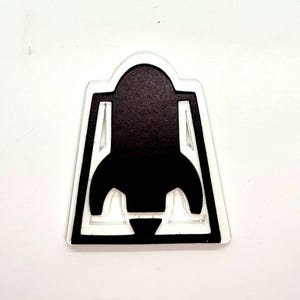 May include: A black rocket ship design on a clear acrylic background. The rocket is set against a black backdrop within a rectangular shape with rounded top edges. The design is on a white surface.