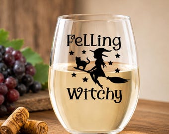 May include: A clear stemless wine glass featuring a black graphic print of a witch riding a broomstick with a cat and stars. The text "FeLling Witchy" is printed in black lettering above and below the graphic on the front of the glass.