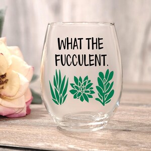May include: A clear stemless wine glass with the text "WHAT THE FUCCULENT." in black. Below the text are three green succulent plant designs. The glass is on a wooden surface with a blurred flower in the background.