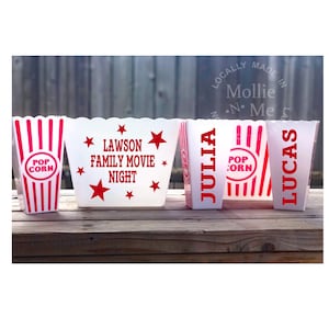 May include: Several white popcorn containers with red stripes and text. One says "POP CORN", another reads "LAWSON FAMILY MOVIE NIGHT", and two others have names. The containers are displayed on a wooden surface.