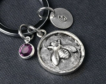 Può includere: Portachiavi d'argento con un ciondolo a forma di ape, un ciondolo di pietra preziosa viola e un ciondolo d'argento con la lettera "G" sopra.