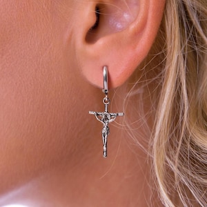 Könnte beinhalten: Silberne Creolen mit einem silbernen Kreuzanhänger. Das Kreuz zeigt eine detaillierte Figur von Jesus Christus.