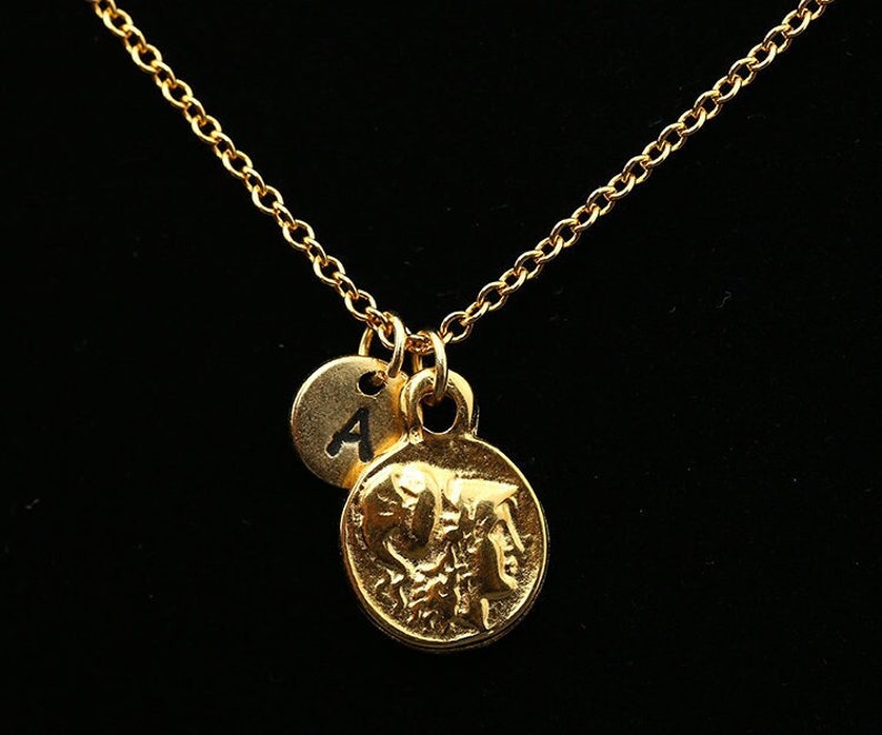 May include: A gold chain necklace with two charms. The larger charm is a round coin with a detailed design. The smaller charm is a round disc with the letter "A" engraved on it.