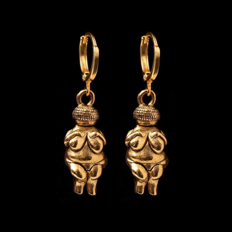 May include: A pair of gold-toned earrings featuring a stylized female figure with a rounded head and a simple, abstract body. The earrings have a hoop design.