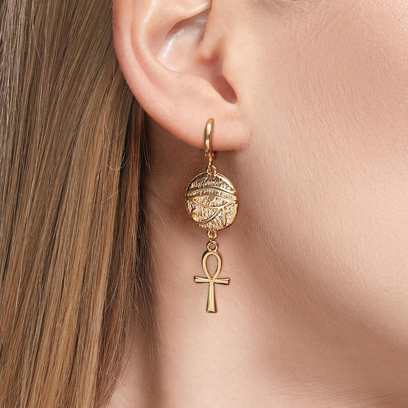 May include: Gold hoop earrings with a round charm featuring an eye design and a dangling ankh symbol.