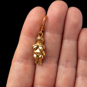 May include: A gold-colored pendant featuring a stylized female figure with arms crossed over her chest. The pendant is attached to a gold-colored chain.