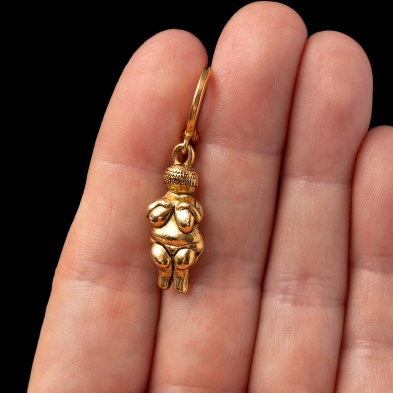 May include: A gold-colored pendant featuring a stylized female figure with arms crossed over her chest. The pendant is attached to a gold-colored chain.