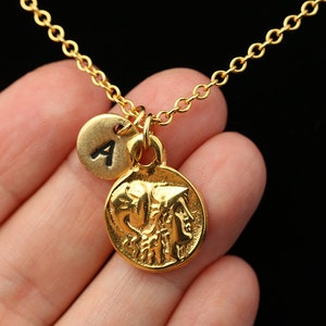May include: Gold necklace with a round coin pendant featuring a profile of a woman's face and a smaller round charm with the letter 'A' on it.