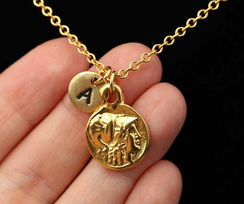 May include: Gold necklace with a round coin pendant featuring a profile of a woman's face and a smaller round charm with the letter 'A' on it.