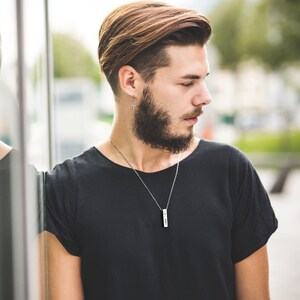 May include: A man with a beard and short brown hair is wearing a black t-shirt with a silver chain necklace and a pendant.