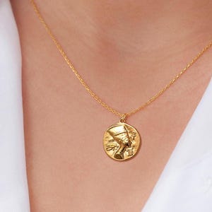 Könnte beinhalten: Eine goldene Kettenkette mit einem runden Anhänger, der ein Profil einer ägyptischen Königin mit Kopfbedeckung zeigt.