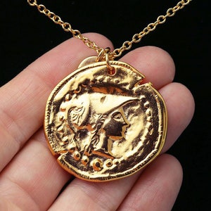 May include: Gold necklace with a large, round coin pendant featuring a detailed profile of a woman wearing a helmet. The coin has a textured surface and a raised rim.