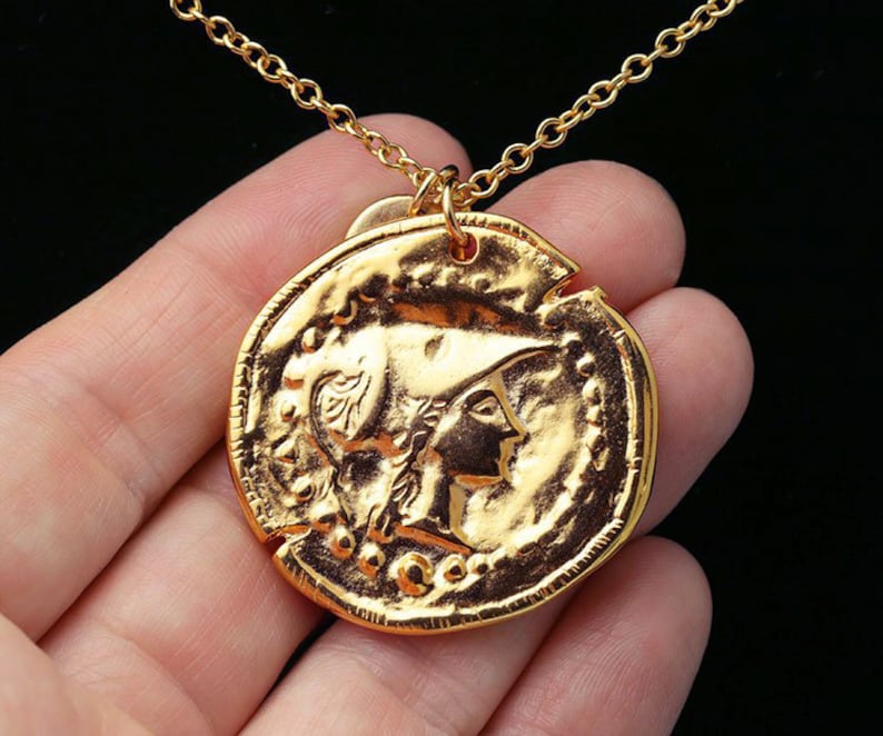 May include: Gold necklace with a large, round coin pendant featuring a detailed profile of a woman wearing a helmet. The coin has a textured surface and a raised rim.