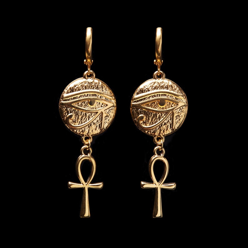 May include: Gold earrings with a textured round pendant featuring the Eye of Horus and a dangling ankh charm.
