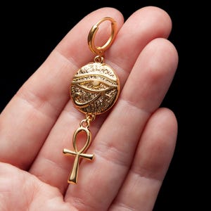 May include: Gold-tone earring with a round pendant featuring the Eye of Horus and a dangling ankh symbol.