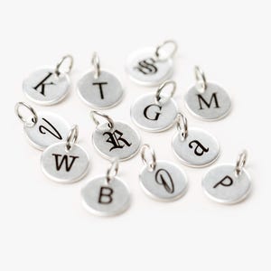May include: A collection of silver charms with different letters engraved on them. The letters include K, T, S, G, M, a, W, R, D, B, and P.