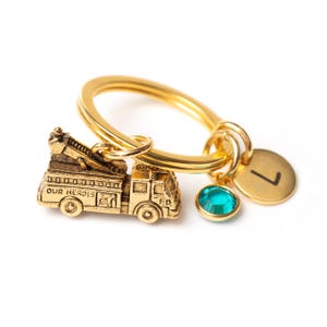 May include: Gold-tone keychain featuring a miniature fire truck charm with the words "Our Heroes" and "FD" on the side, a teal blue crystal charm, and a gold-tone charm with the letter "L" on it.