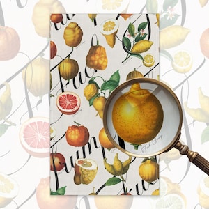 May include: A white rectangular towel with a botanical illustration of citrus fruits in yellow, orange, and red. The towel has the words "Fuck Trumps" in a black script font. A magnifying glass is in the foreground.