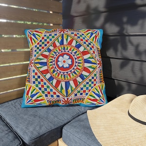May include: Square cushion with a vibrant, colourful geometric design. The cushion features a central floral motif and a bright blue border. The design incorporates red, yellow, blue, and green elements.