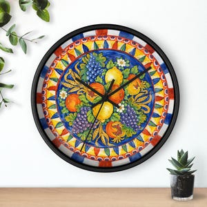 Op de afbeelding: Een ronde wandklok met een zwart frame. De wijzerplaat toont een kleurrijke illustratie van fruit, waaronder druiven, citroenen en granaatappels. Het ontwerp is geplaatst tegen een blauwe achtergrond met een decoratieve rand.