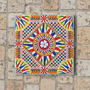 May include: Square, colorful tile with a vibrant, symmetrical design. The pattern features a central floral motif surrounded by geometric shapes in red, blue, yellow, and green. The tile is set against a stone background.