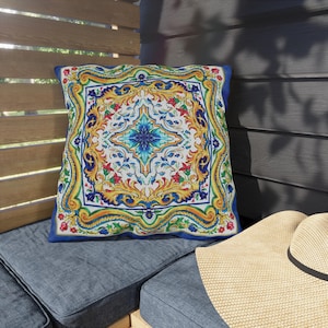 May include: Square decorative pillow with a vibrant, ornate design. The pillow features a central blue starburst pattern surrounded by a symmetrical arrangement of yellow, red, and green floral motifs on a white background, framed by a blue border.