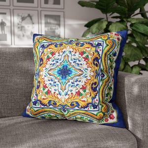 May include: Square cushion with a colourful, ornate design. The cushion features a central blue starburst pattern surrounded by a detailed floral and scrollwork border in yellow, red, and green on a white background. It has a solid navy blue border.