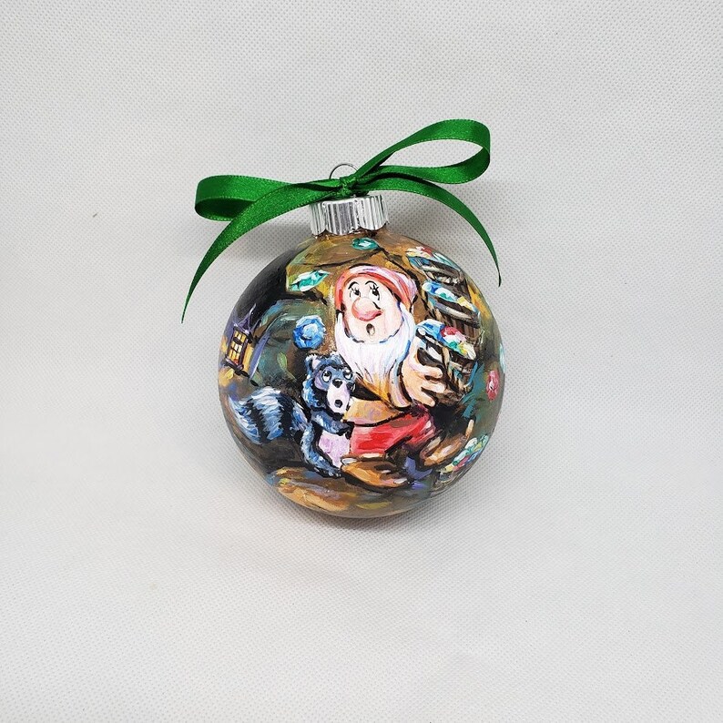 May include: A hand-painted Christmas bauble featuring a cartoon dwarf holding a bag of gems. The dwarf is wearing a red and white outfit and has a surprised expression on his face. A small black and white animal is standing next to him. The bauble is decorated with a green ribbon.