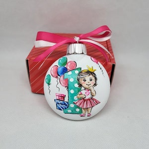 May include: A white glass bauble with a hand-painted design of a girl wearing a pink dress and a crown, holding a heart. The girl is standing in front of a large green number one with white polka dots. There are colourful balloons and presents in the background.