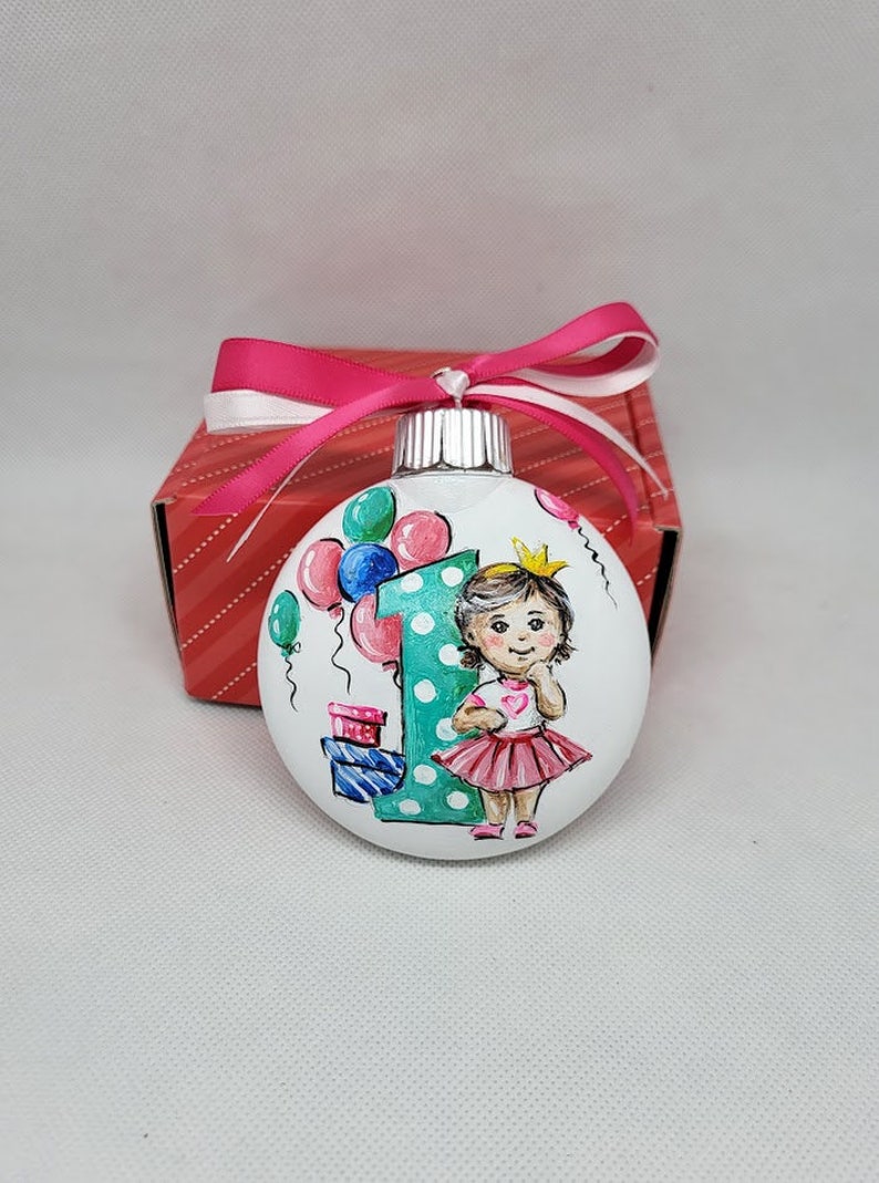 May include: A white glass bauble with a hand-painted design of a girl wearing a pink dress and a crown, holding a heart. The girl is standing in front of a large green number one with white polka dots. There are colourful balloons and presents in the background.