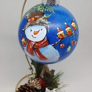 May include: A hand-painted Christmas bauble featuring a snowman with a red scarf and hat, surrounded by red robins on a blue background. The bauble is on a gold stand with pine cones and greenery.