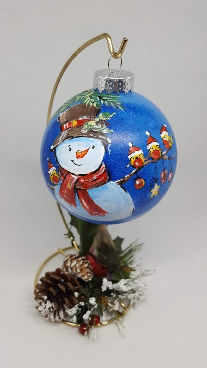 May include: A hand-painted Christmas bauble featuring a snowman with a red scarf and hat, surrounded by red robins on a blue background. The bauble is on a gold stand with pine cones and greenery.