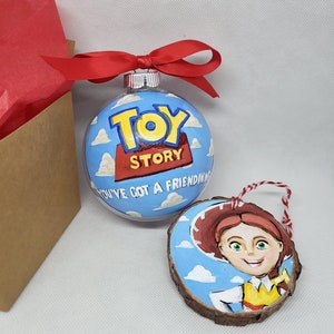 May include: A set of two Christmas decorations featuring characters from the film Toy Story. The first decoration is a clear glass bauble with a blue background and white clouds. The words "Toy Story" are painted in red and yellow on the decoration. The second decoration is a wooden circle with a blue background and white clouds. The character Jessie from Toy Story is painted on the decoration.