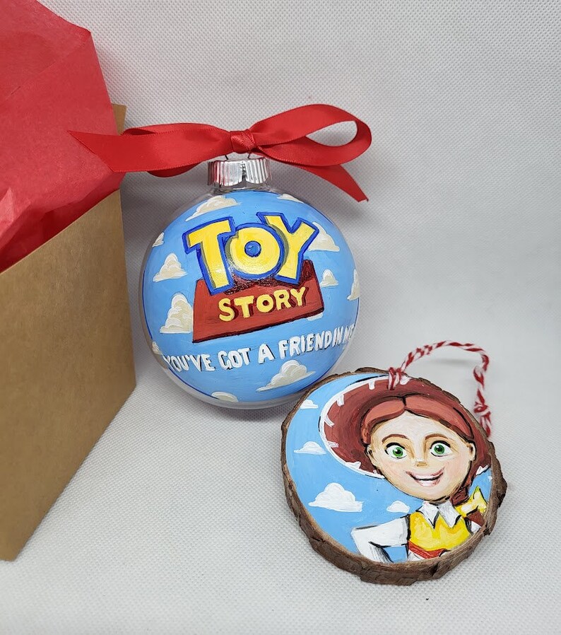 May include: A set of two Christmas decorations featuring characters from the film Toy Story. The first decoration is a clear glass bauble with a blue background and white clouds. The words "Toy Story" are painted in red and yellow on the decoration. The second decoration is a wooden circle with a blue background and white clouds. The character Jessie from Toy Story is painted on the decoration.