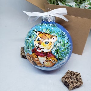 May include: A blue glass Christmas bauble with a hand-painted image of a tiger cub wearing a red scarf and surrounded by green pine branches and white snowflakes.